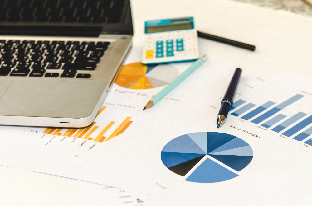 Laptop, calculator, pen and financial reports with bar and pie charts spread across a desk.
